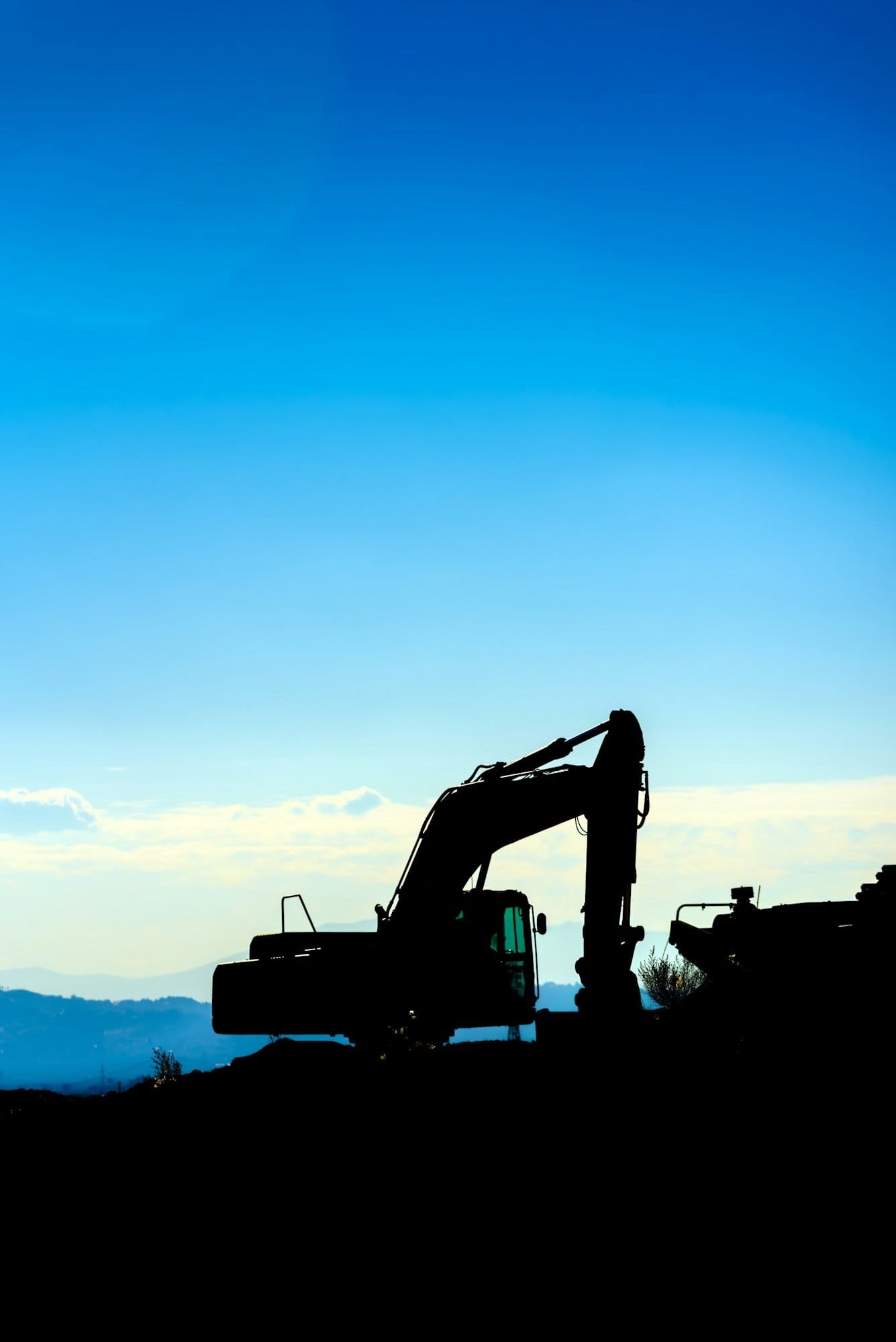 Backlit silhouette of an excavator on top of a hill with unfocused sky background.